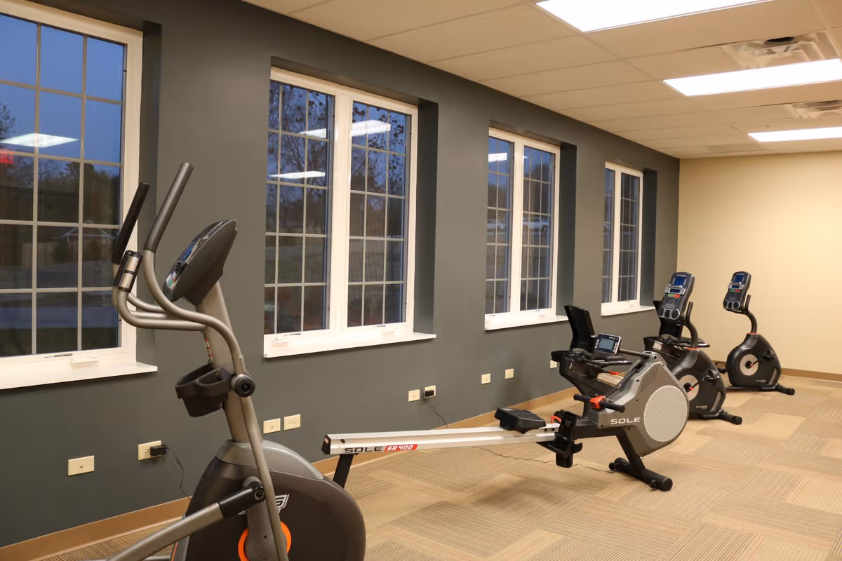 A fitness room with exercise equipment including an elliptical machine, a rowing machine, and two stationary bikes. The room has large windows along one wall, beige carpeted floor, and a combination of dark green and beige walls with a drop ceiling and fluorescent lighting.