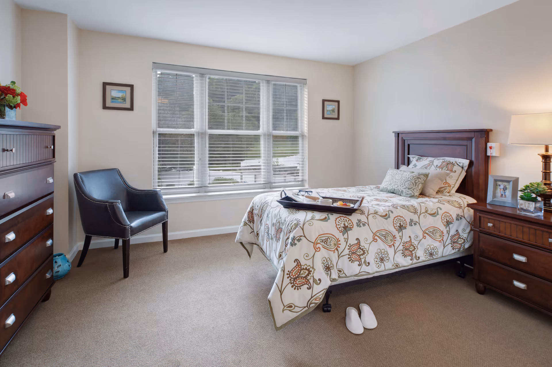Sunlit bedroom with a single bed, wooden nightstand and dresser, an upholstered chair, and a large window.