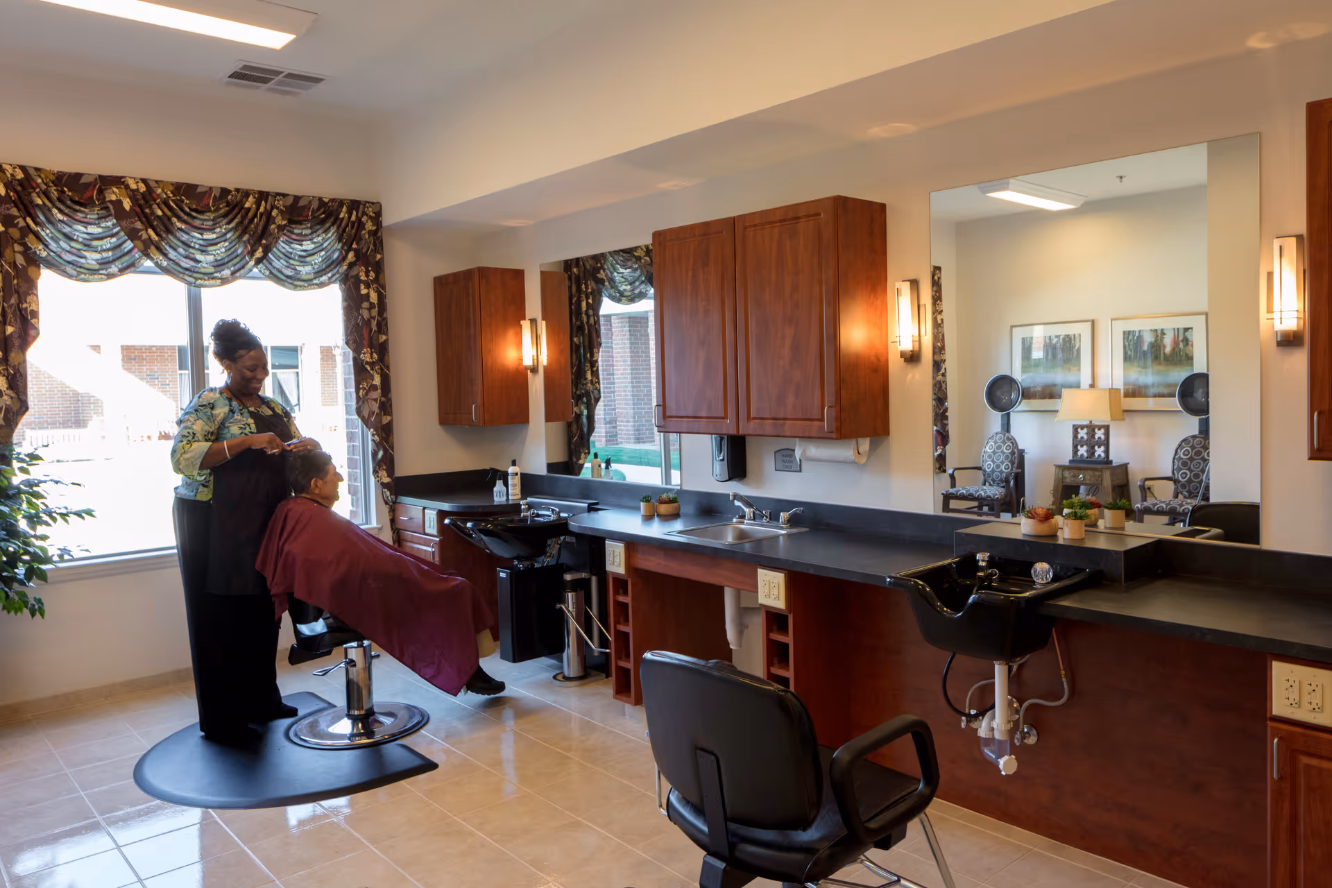 A hair salon area inside a senior living facility with a stylist cutting a seated woman's hair. The room has large mirrors, wooden cabinets, a sink, and salon chairs. A window with floral curtains lets in natural light, and a seating area with chairs and a lamp is reflected in the mirror.