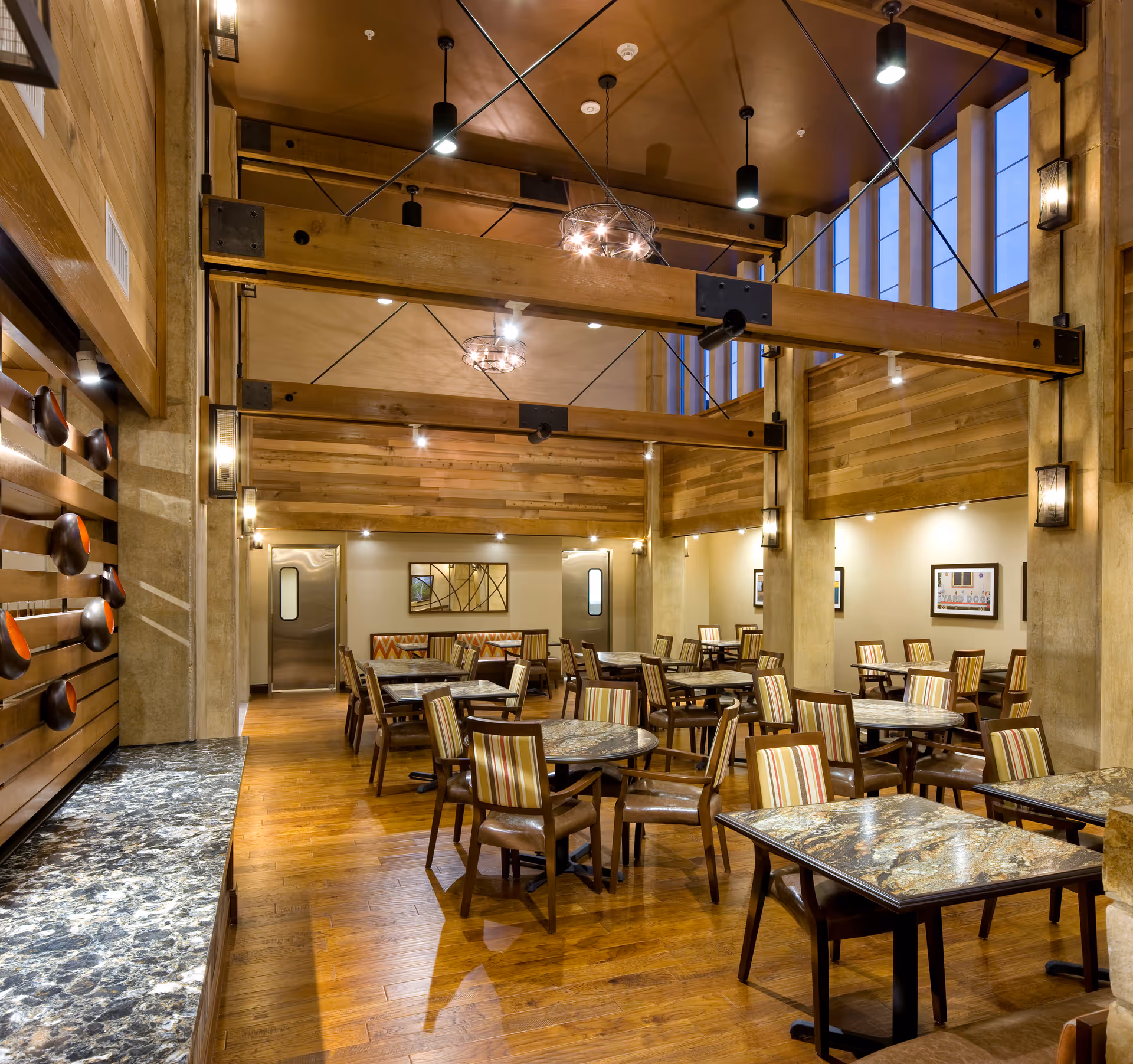 Spacious, warmly lit dining room featuring exposed wooden beams, marble-topped tables and striped upholstered chairs.