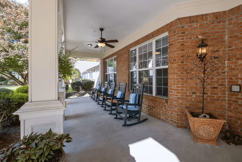 Covered outdoor porch area with a row of black rocking chairs, each with a blue and white striped cushion, against a brick wall with large windows. There is a ceiling fan with a light fixture above and potted plants on the concrete floor. The porch overlooks a garden with bushes and trees.