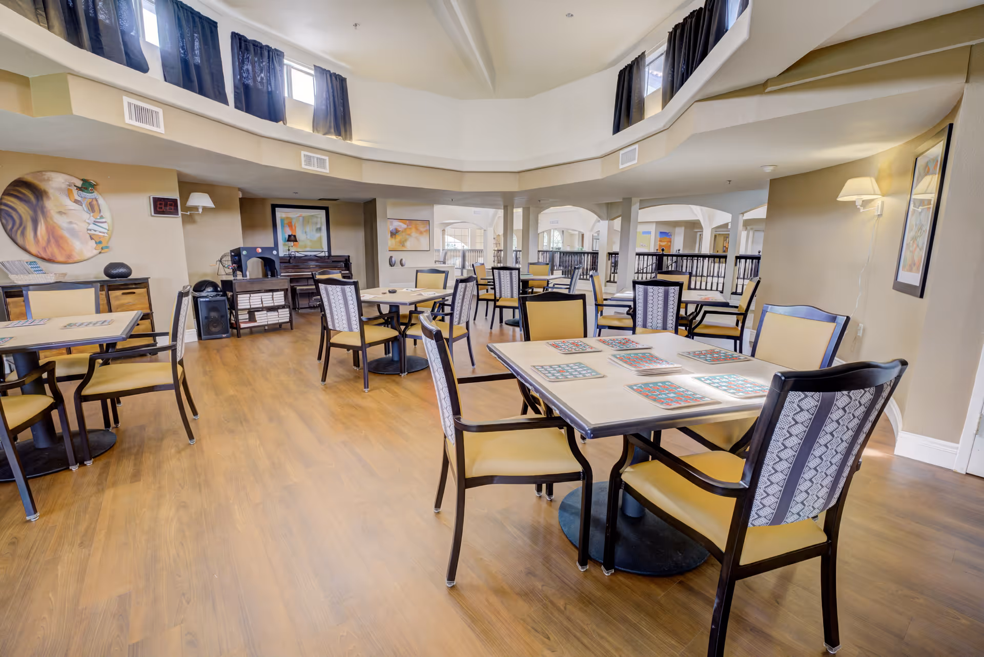 A spacious dining room with multiple tables and chairs arranged neatly on a wooden floor. The room features high ceilings with windows near the top letting in natural light, beige walls adorned with framed artwork, and soft lighting from wall-mounted lamps. The tables have placemats and the overall atmosphere is clean and inviting.