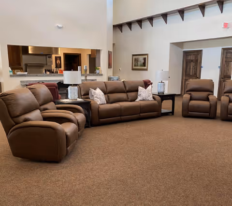 Spacious communal living room with brown leather reclining sofas and chairs arranged around end tables and lamps, with a kitchen pass-through in the background.
