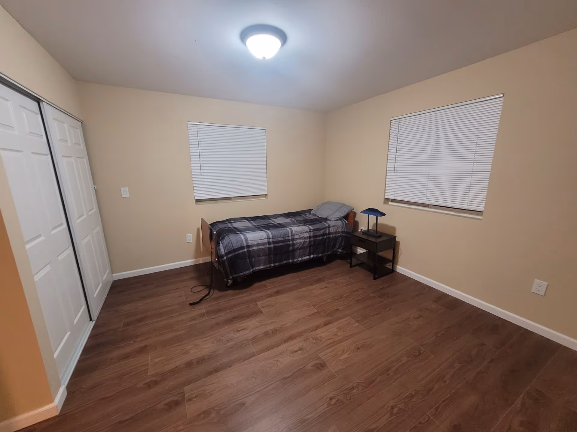 A simple bedroom with beige walls and wood flooring. The room contains a single bed with a gray and black plaid blanket and a gray pillow. Next to the bed is a small black nightstand with a blue lamp. There are two windows with closed white blinds and a closet with white sliding doors.