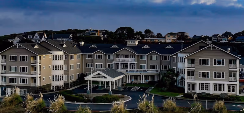 Front exterior of a multi-story senior living building with balconies, a covered entrance, circular driveway, and landscaping.