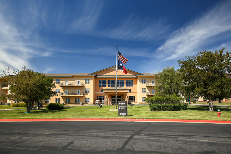 Front exterior view of The Craig Senior Living facility, a three-story building with balconies, a flagpole displaying the American and Texas flags, surrounded by green lawns and trees under a blue sky with some clouds.
