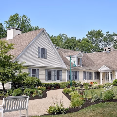 Exterior view of a single-story building with beige siding and a brown shingled roof, surrounded by well-maintained landscaping including bushes, flowers, and a tree. A curved pathway leads to the entrance, and a white bench is placed near the path. The sky is clear and blue.