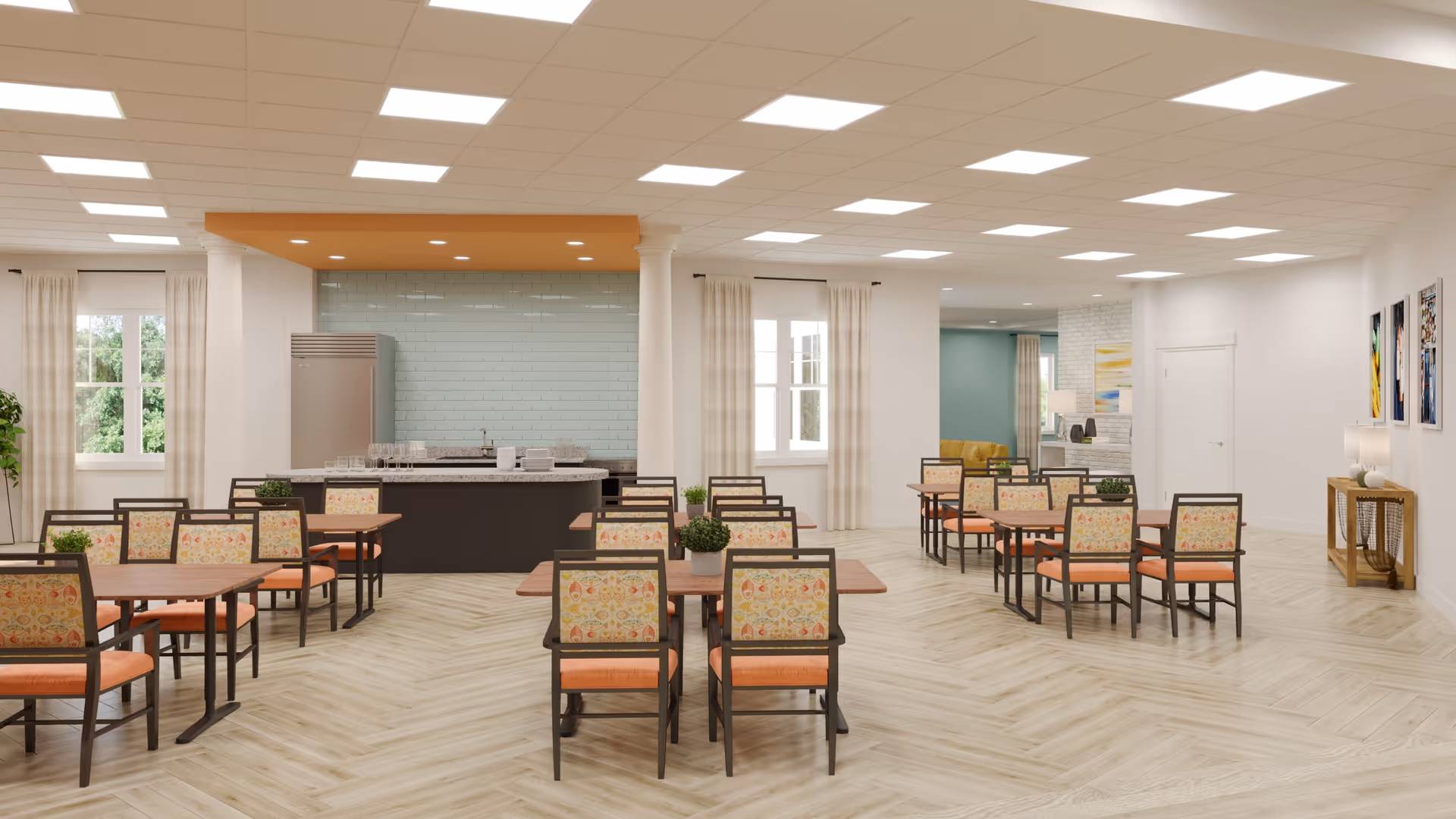 A spacious dining room with multiple wooden tables and chairs featuring floral upholstery and orange cushions. The room has large windows with light curtains, a kitchen area with a refrigerator and sink, light wood flooring, and bright ceiling lights. There are some small potted plants on the tables and colorful artwork on the walls.