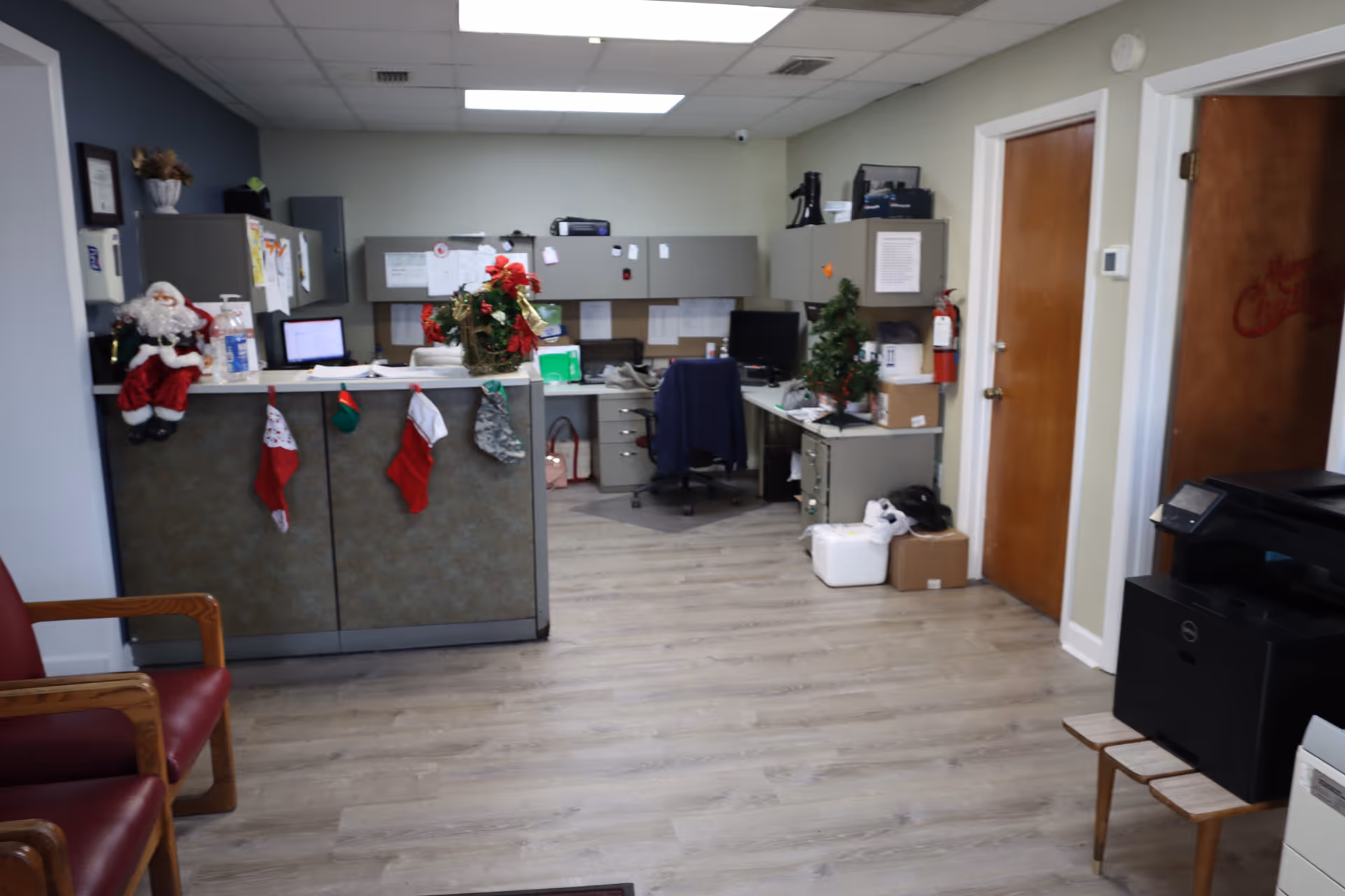 An office area with a reception desk decorated with Christmas stockings and a Santa Claus figure. Behind the desk are computer workstations, filing cabinets, and office supplies. The floor is light wood, and there are two wooden doors on the right side of the room. A red chair is partially visible on the left, and a printer is on the right.