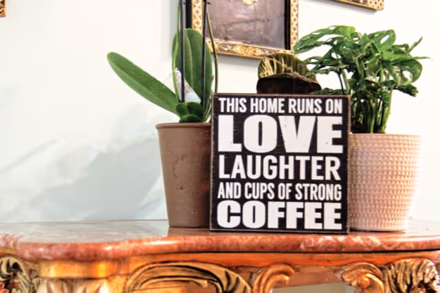 Ornate wooden console topped with two potted plants and a decorative sign reading "This home runs on love, laughter and cups of strong coffee".