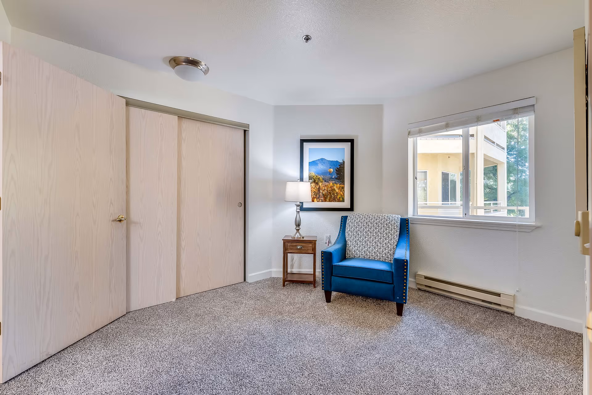 Carpeted room with a blue upholstered armchair beside a wooden side table and lamp, a framed picture on the wall, sliding closet doors, and a window.