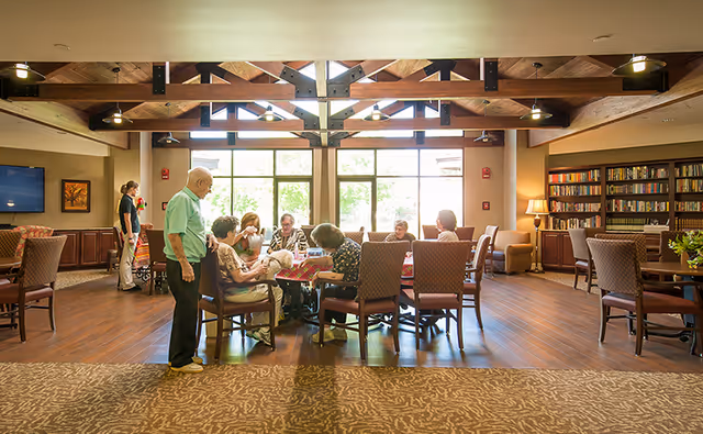 Seniors and a staff member gathered around a table playing cards in a spacious assisted-living common room with exposed wood beams, bookshelves, and large windows.
