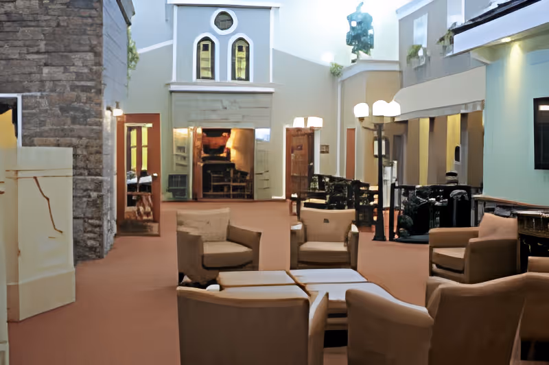 A spacious living room area in a senior living facility with several beige armchairs arranged around a central coffee table. The room features a high ceiling with a decorative wall resembling a house facade, including windows and a fireplace. There are lamps providing warm lighting and a stone wall on the left side.