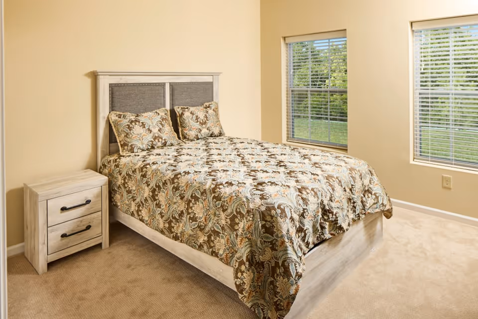 A bedroom with a bed featuring a floral patterned bedspread and two matching pillows. The bed has a light wood frame with a padded headboard. Next to the bed is a small matching nightstand with two drawers. The room has beige walls, beige carpet, and two windows with white blinds showing green trees outside.