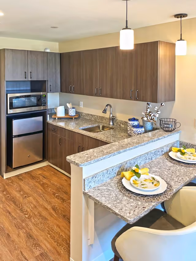 A modern kitchen area with wooden cabinets, granite countertops, a stainless steel microwave and mini refrigerator. There is a small sink with a faucet, decorative containers, and a coffee mug on the counter. In the foreground, a granite breakfast bar is set with two plates, yellow napkins, and small floral decorations, accompanied by two cushioned chairs. The floor is wood, and two pendant lights hang from the ceiling.