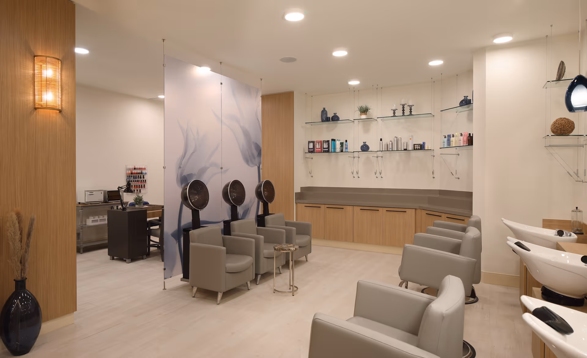 A modern salon area with multiple gray salon chairs, hair drying stations with hooded dryers, and washing sinks. The room features light wood cabinetry, glass shelves with hair care products, and soft lighting. A decorative panel with a floral design separates part of the space.