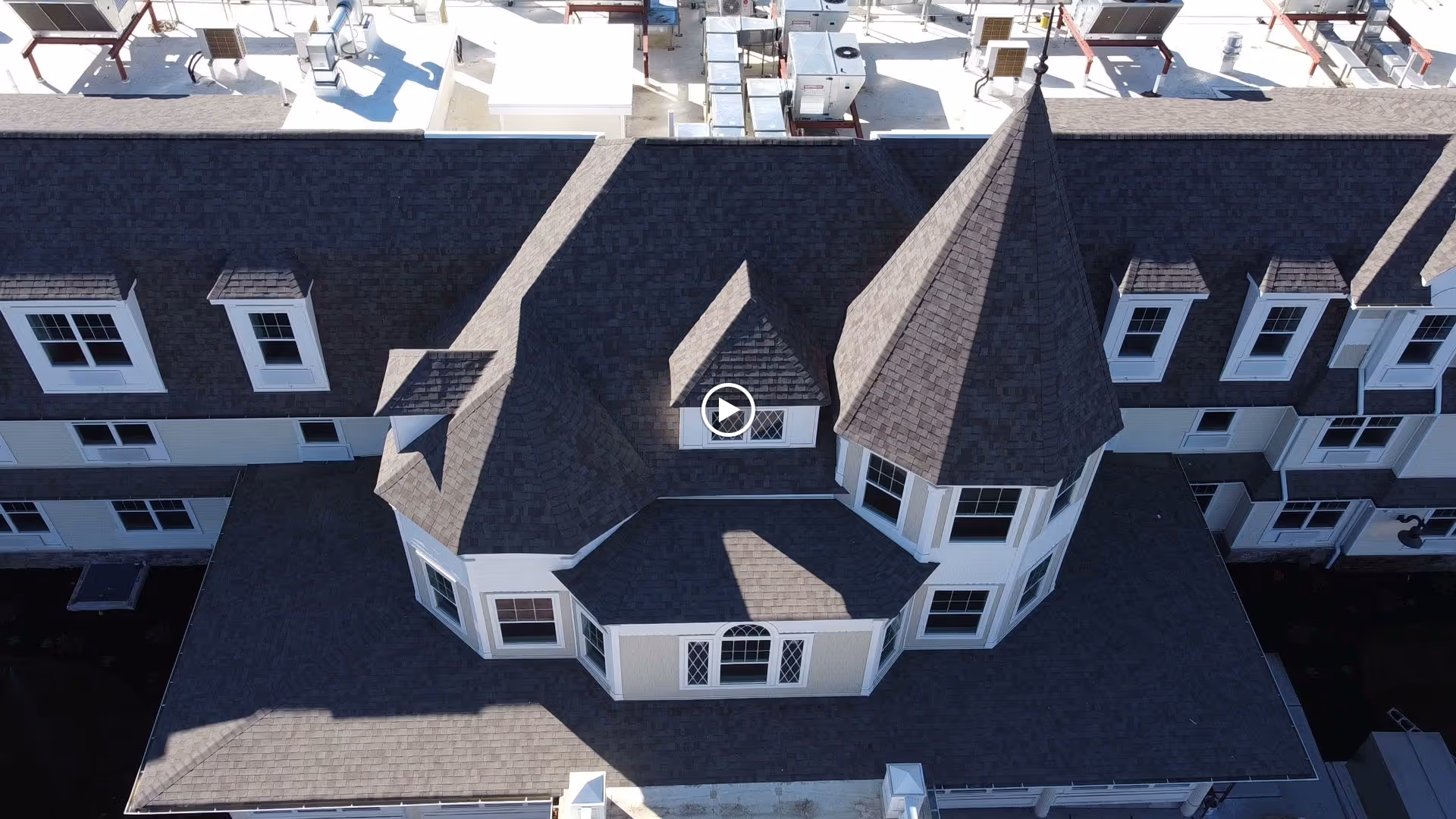 Aerial view of a building roof with multiple gables and a prominent turret-like structure, featuring dark shingles and several white-framed windows.