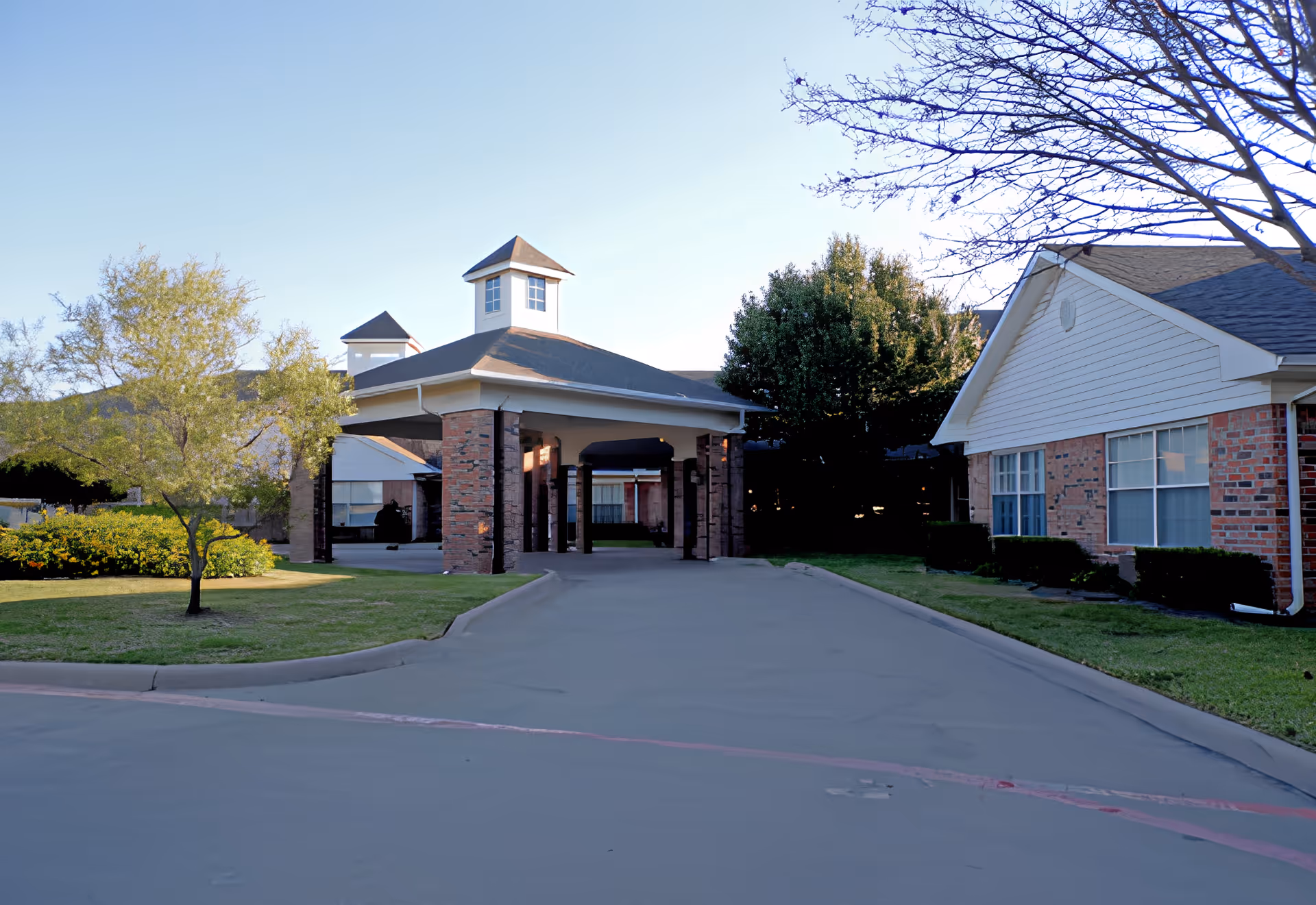 Exterior view of Crestwood Health and Rehabilitation Center showing a covered entrance with brick pillars, surrounded by green lawns, trees, and bushes under a clear sky.