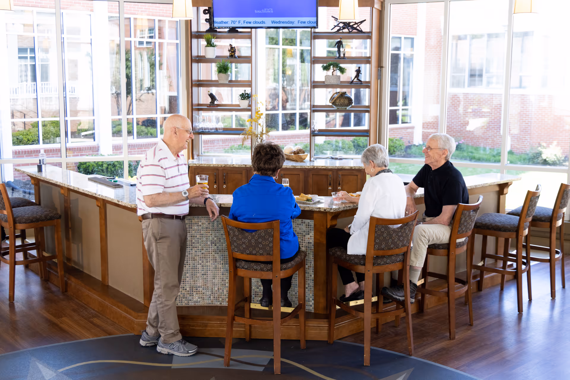 Four senior residents socializing around a counter bar in a bright common dining area with large windows.