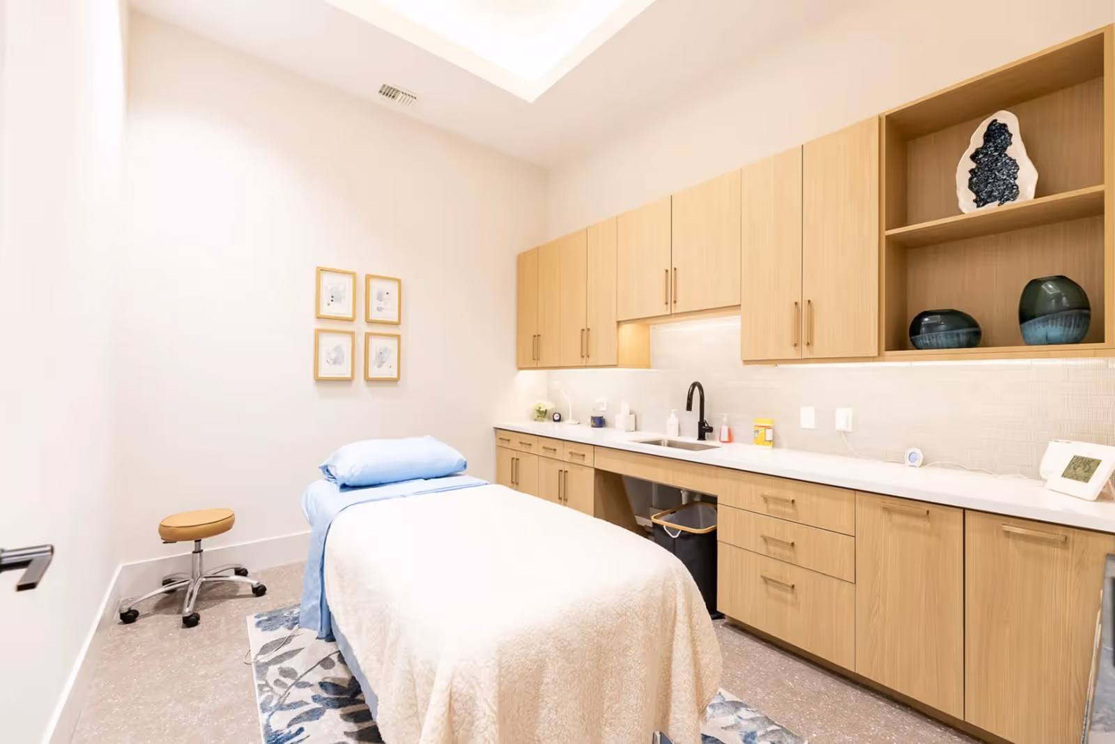 A clean, well-lit treatment room with a massage or examination table covered with a white blanket and blue sheets. The room features light wood cabinetry with a sink, decorative vases, and framed artwork on the wall. There is a small rolling stool next to the table and a patterned rug underneath.