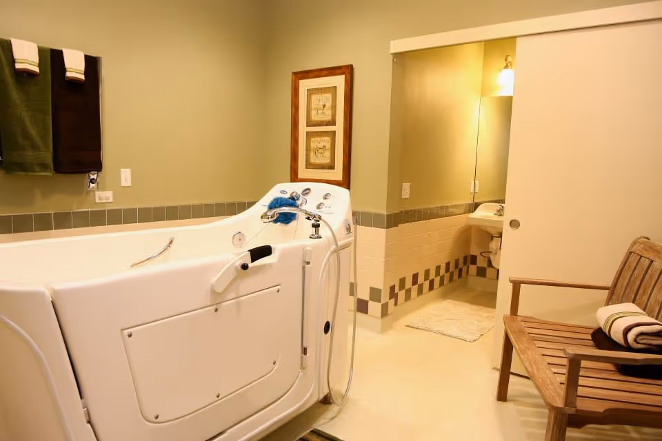 A bathroom featuring a large white walk-in bathtub with handrails and a handheld showerhead. On the wall above the tub, there are two framed pictures and two towels hanging on a rack. To the right, there is a wooden bench with a rolled towel on it. In the background, a sink with a mirror and light fixture is visible through an open doorway.