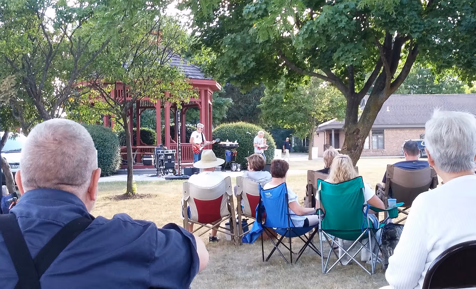 A group of people sitting on lawn chairs outdoors, watching a live music performance near a red gazebo surrounded by trees and bushes in a grassy area.