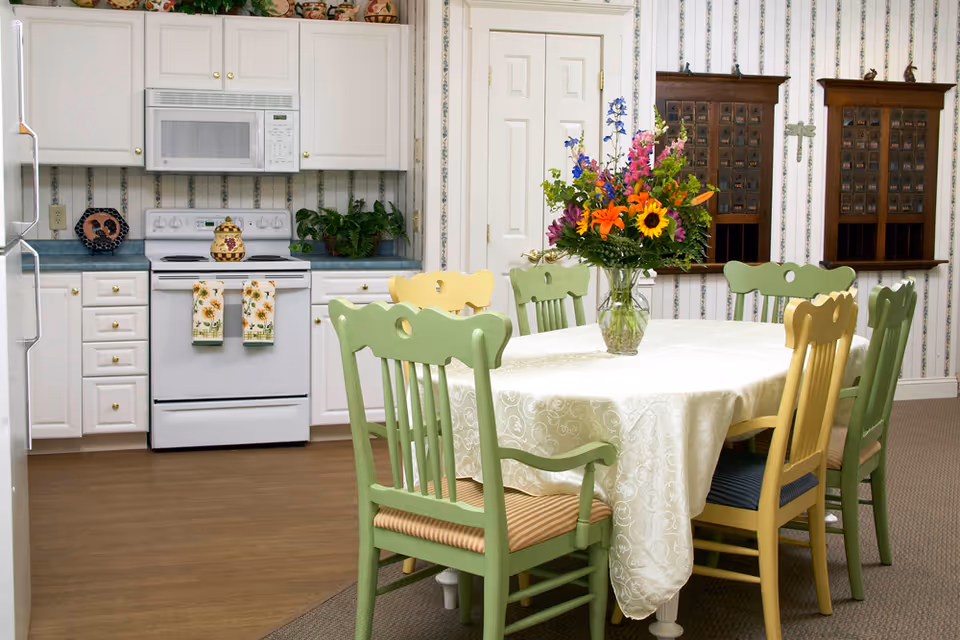 A cozy kitchen and dining area featuring white cabinets, a white stove with a microwave above it, and a dining table covered with a white tablecloth. The table is surrounded by green and yellow wooden chairs, and a vase with colorful flowers sits in the center. The walls have striped wallpaper with two wooden mail organizers mounted on them.