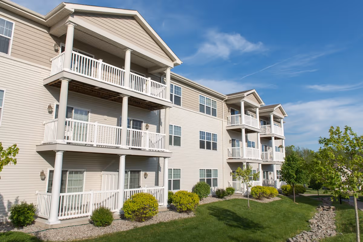 Exterior view of a three-story residential building with beige siding, white balconies, and columns. The building is surrounded by green grass, small bushes, and young trees under a blue sky with some clouds.