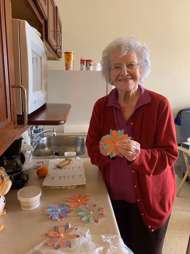 An elderly woman smiles in a small kitchenette holding colorful paper flower crafts with several more on the counter.
