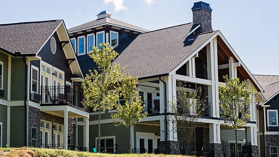 Exterior view of a multi-story senior living facility building with green siding, stone accents, balconies, and several young trees in front under a clear blue sky.