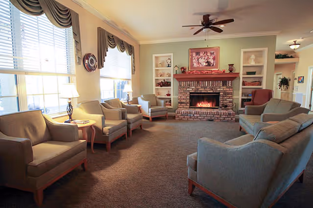 A cozy living room with beige armchairs and sofas arranged around a brick fireplace with a fire burning. The room has large windows with decorative valances, built-in white shelves flanking the fireplace, and a ceiling fan above.