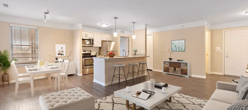 A spacious and modern open-concept living area in a senior living facility apartment. The room features a kitchen with stainless steel appliances, a breakfast bar with three stools, a dining area with a white table and chairs, and a living room with a sofa, ottoman, and coffee table on a patterned rug. The walls are painted beige, and there are decorative plants and artwork. The flooring is wood-style laminate.