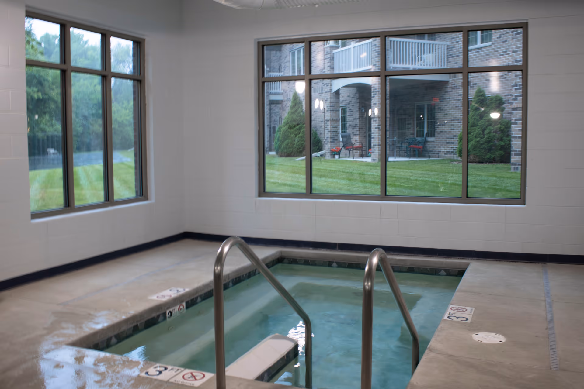 Indoor small pool or hot tub with metal handrails, surrounded by concrete floor and large windows showing green grass and a brick building outside.