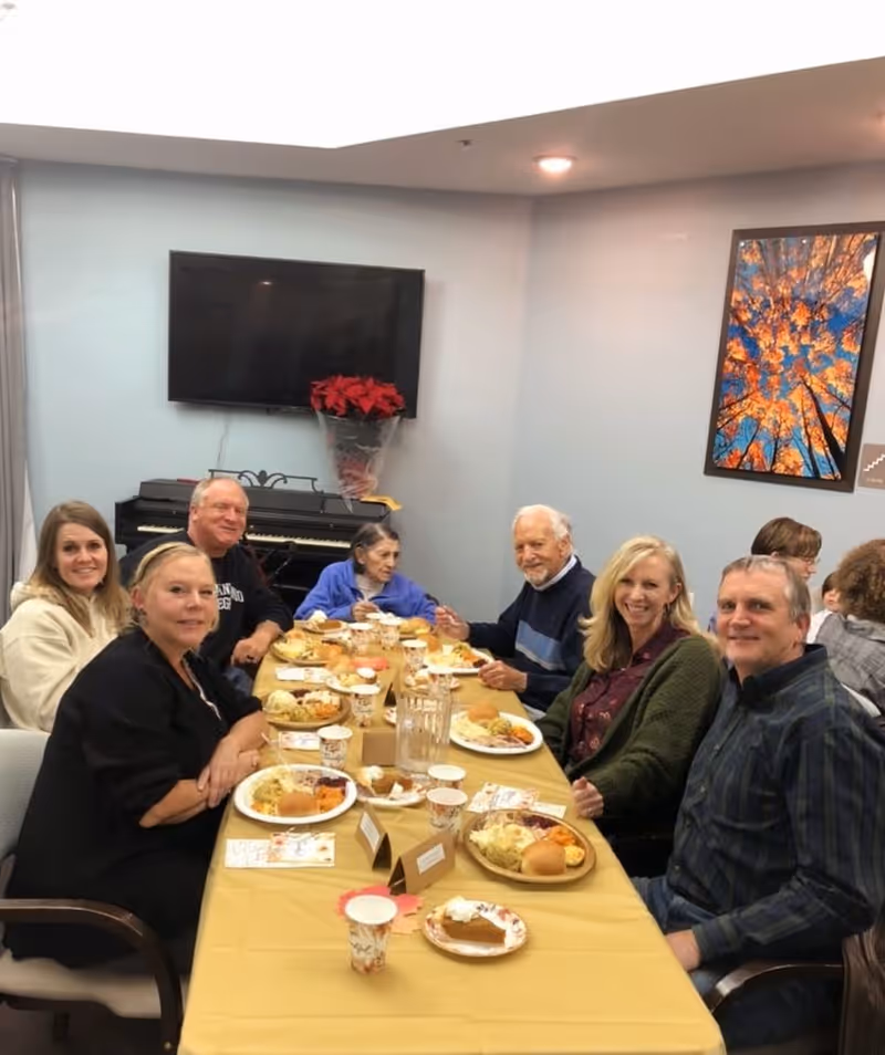 A group of seven people, including elderly individuals and caregivers, sitting around a table enjoying a meal together in a well-lit room. The table is covered with a yellow tablecloth and has plates of food, cups, and a pitcher of water. A piano with a poinsettia plant on top and a large flat-screen TV are visible on the wall behind them. A colorful framed picture of autumn trees is hung on the adjacent wall.