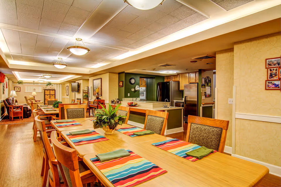 Spacious communal dining room with a long wooden table set with colorful placemats and a potted centerpiece, chairs, an open kitchen, and a seating area in the background.