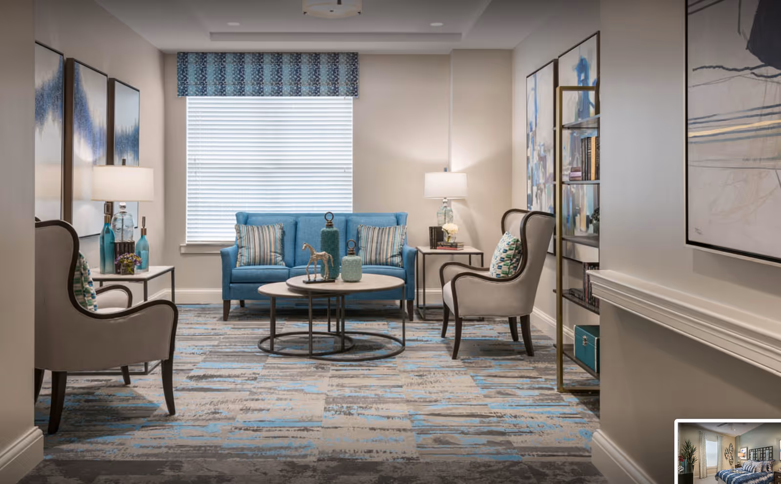 A cozy living room with a blue sofa, two gray armchairs with patterned cushions, two round nested coffee tables with decorative items, two side tables with lamps, abstract artwork on the walls, and a window with blinds and a blue valance.