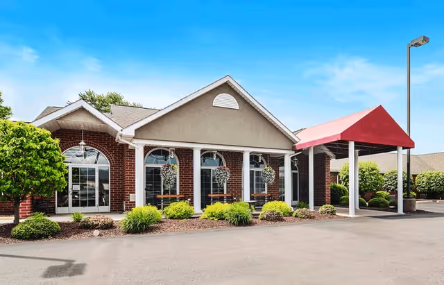 Exterior view of a single-story brick building with a beige gable roof and white columns supporting a red canopy over the entrance. There are hanging flower baskets along the front windows and neatly trimmed bushes and greenery in front of the building. The sky is clear and blue.