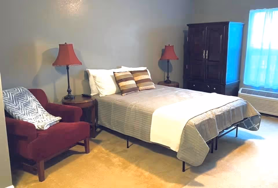 A bedroom with a neatly made bed featuring striped bedding and two decorative pillows. On each side of the bed are wooden nightstands with matching red lamps. To the left of the bed is a red armchair with a patterned throw blanket draped over it. On the right side of the room is a dark wooden cabinet and a window with sheer blue curtains letting in natural light. The floor is carpeted in a light beige color.