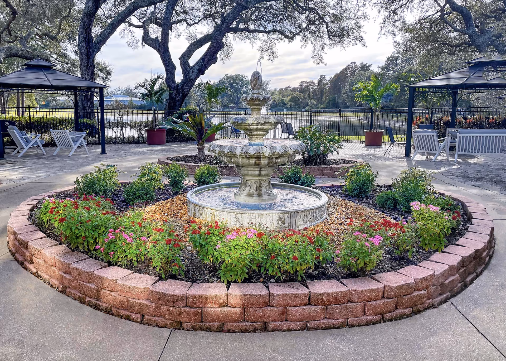 Three-tier stone fountain centered in a circular brick planter filled with flowers, surrounded by seating gazebos and a lakeside courtyard.