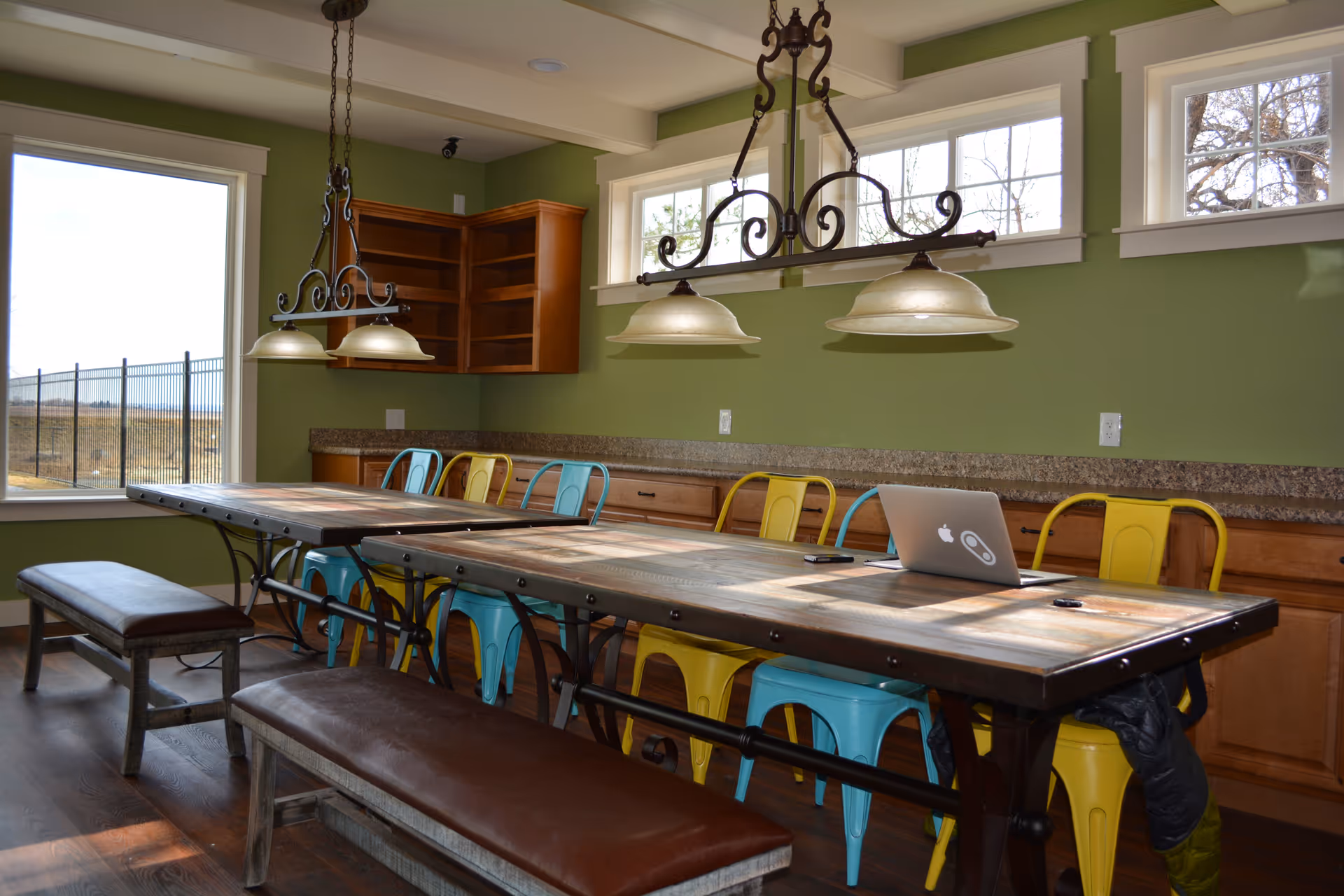 A bright dining room with a long wooden table surrounded by colorful metal chairs in blue and yellow, and two brown cushioned benches. The room has green walls, wooden cabinets, and large windows letting in natural light. Two decorative hanging light fixtures are above the table. A laptop and a phone are placed on the table.
