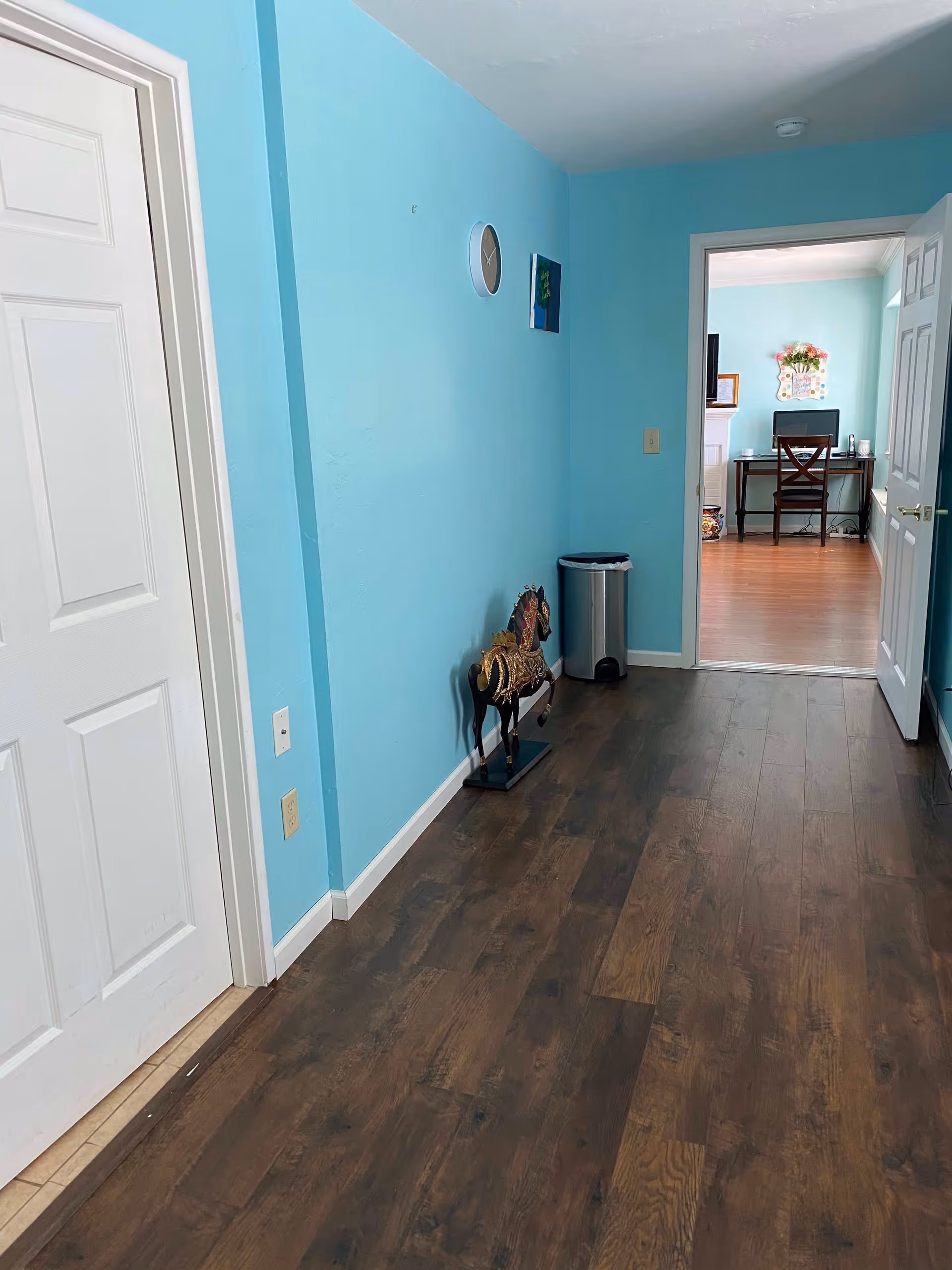 A hallway with light blue walls and dark wood flooring. On the left side, there is a closed white door. On the right side, there is a small decorative horse statue and a silver trash can. At the end of the hallway, an open doorway leads to a room with a desk, chair, and a wall decoration with flowers.