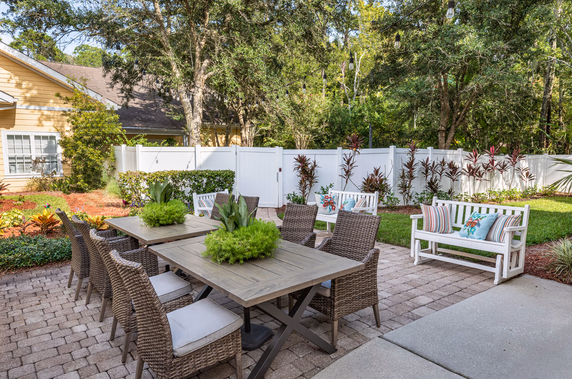 Patio dining area with a wooden table, wicker chairs, and cushioned benches surrounded by plants and a white fence.
