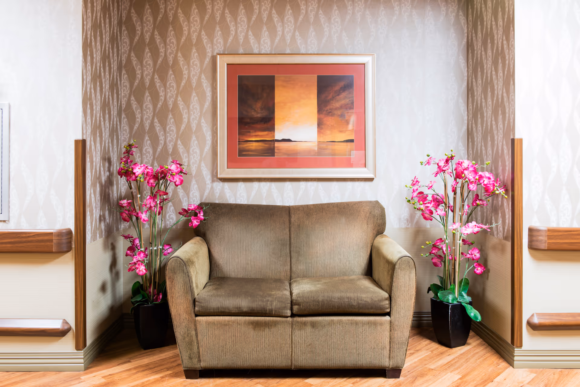 A cozy seating area with a brown two-seater sofa placed against a wall with patterned wallpaper. On either side of the sofa, there are tall black pots with vibrant pink flowers. Above the sofa, a framed artwork featuring a sunset or sunrise scene with warm colors is hung. The floor is wooden, and there are wooden handrails on the walls on both sides.