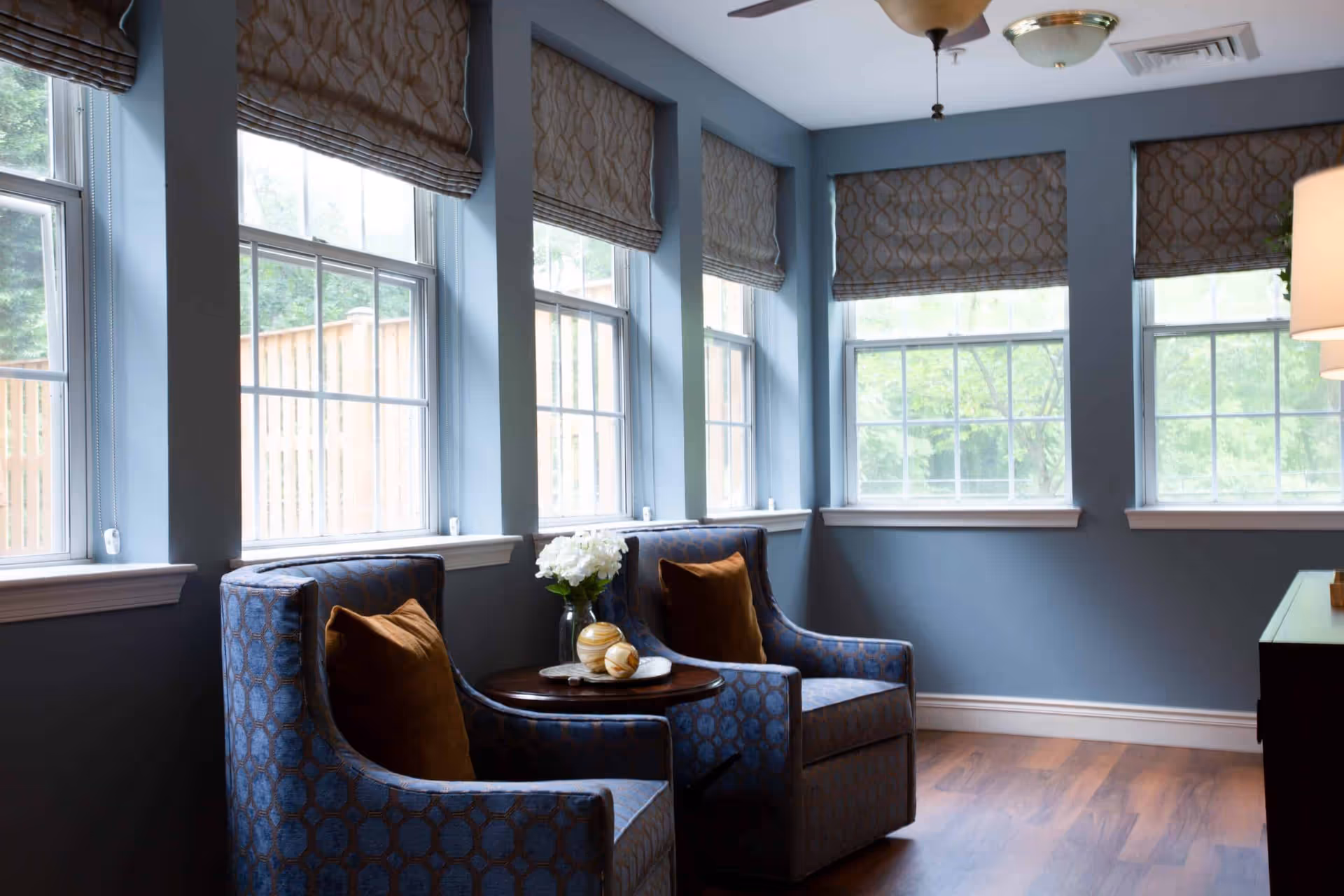 A cozy sitting area with two patterned blue armchairs each with a brown cushion, positioned around a small round wooden table with decorative items and a vase of white flowers. The room has blue walls, multiple large windows with patterned Roman shades, and wooden flooring.