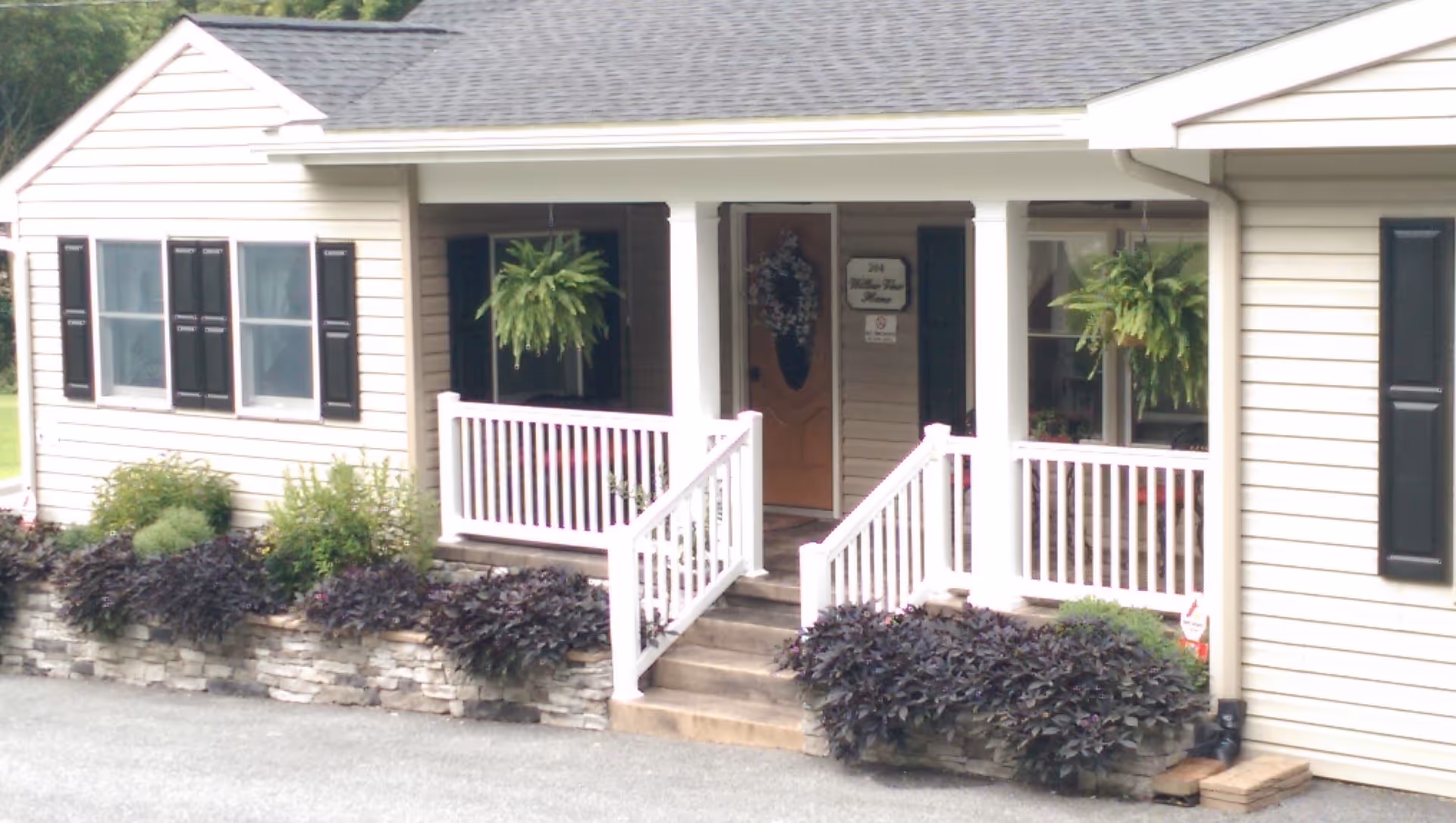 Front exterior view of a single-story building with beige siding, black shutters, and a gray shingled roof. There is a small porch with white railings and steps leading up to a wooden door decorated with a wreath. Two hanging green plants are visible on the porch, and there are landscaped flower beds with various plants along the front of the building.