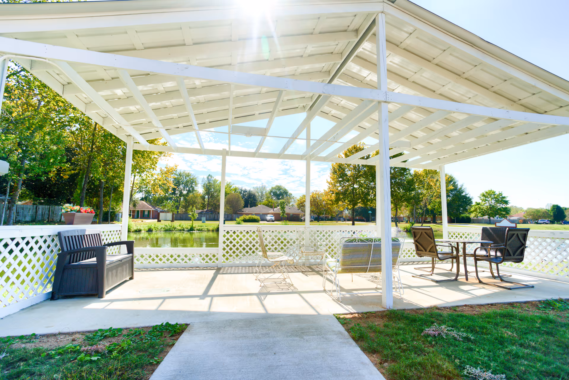Outdoor covered patio area with white lattice railing overlooking a pond. The patio has a white roof structure and contains various seating options including a black bench, metal chairs, a cushioned bench, and a table with chairs. Trees and houses are visible in the background under a clear sky.