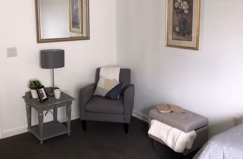 A cozy corner of a bedroom featuring a gray armchair with a decorative pillow and a white throw draped over the back. Next to the chair is a small gray side table with a lamp, a small plant, a framed photo, and a cup. A gray cushioned ottoman with a white blanket and an open book rests nearby. Two framed floral artworks hang on the white walls.