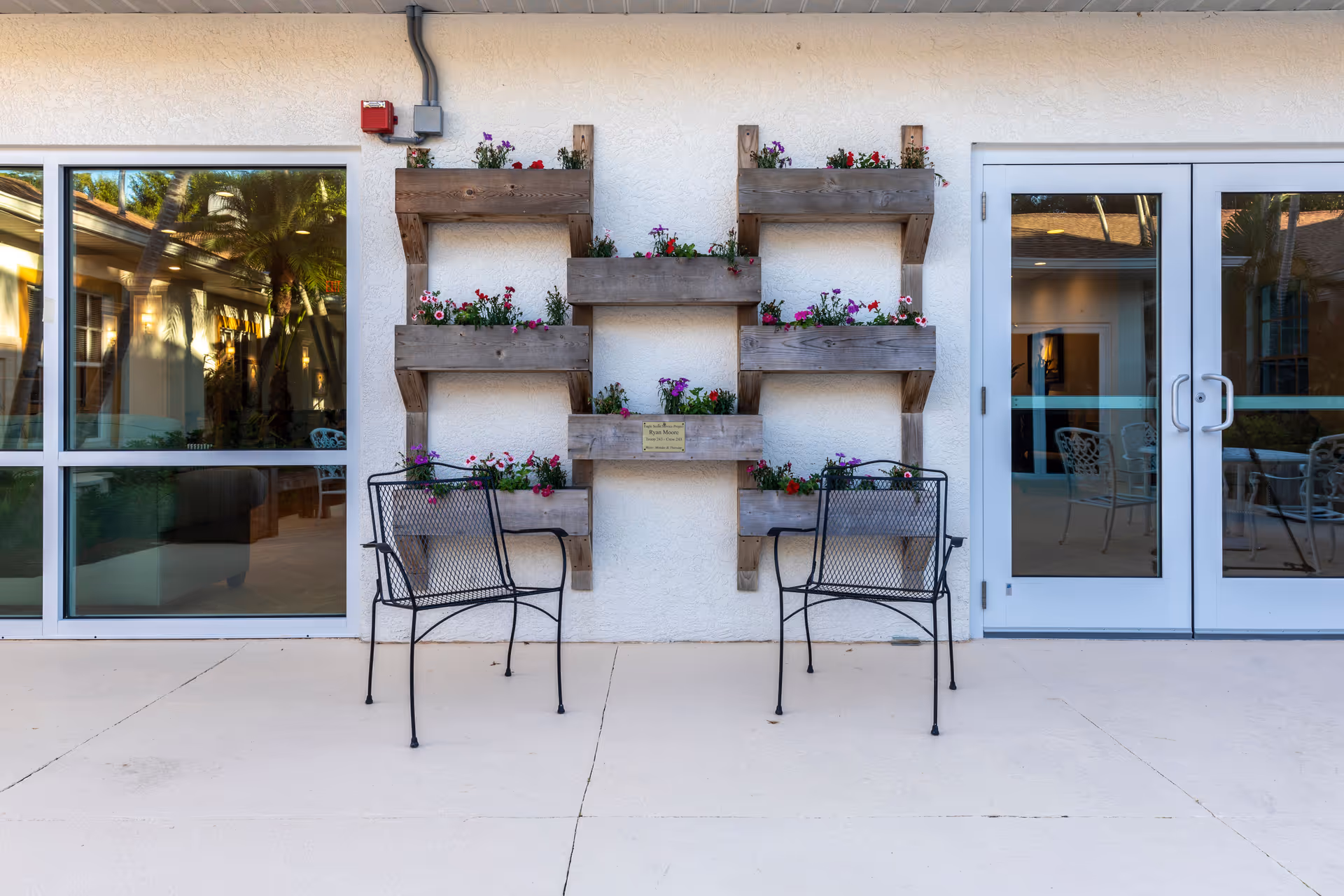 Two black metal chairs sit under a wooden wall planter filled with flowers between windows and glass doors on an exterior patio.