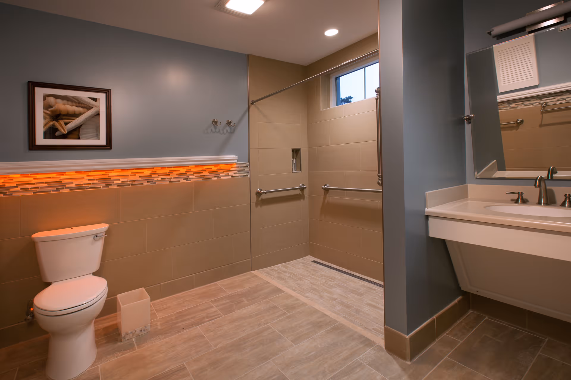 A modern bathroom with a toilet, a walk-in shower with grab bars, a large mirror above a sink with a countertop, and a framed picture on the wall. The walls are painted blue and beige tiles cover the lower half. There is a small window in the shower area and recessed lighting on the ceiling.