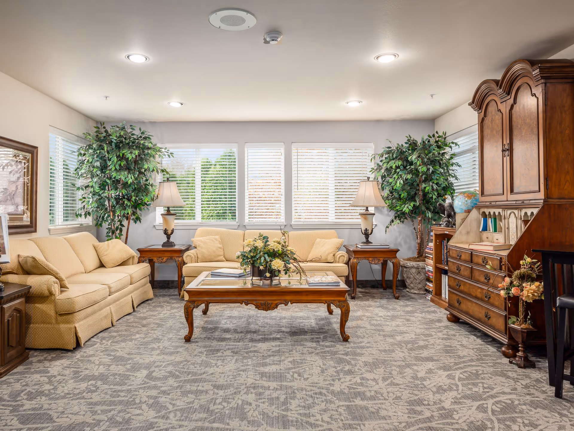 A well-lit living room with two beige sofas, a wooden coffee table with books and a flower arrangement, two side tables each with a lamp, two large potted plants, a wooden cabinet with drawers and shelves, and large windows with white blinds letting in natural light.